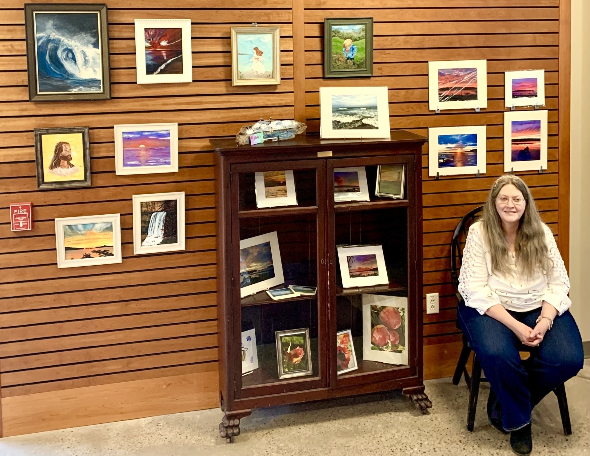 Artist Cathy Stewart sitting on a chair in front of the display of her art work on the wall at Mendon Public Library.