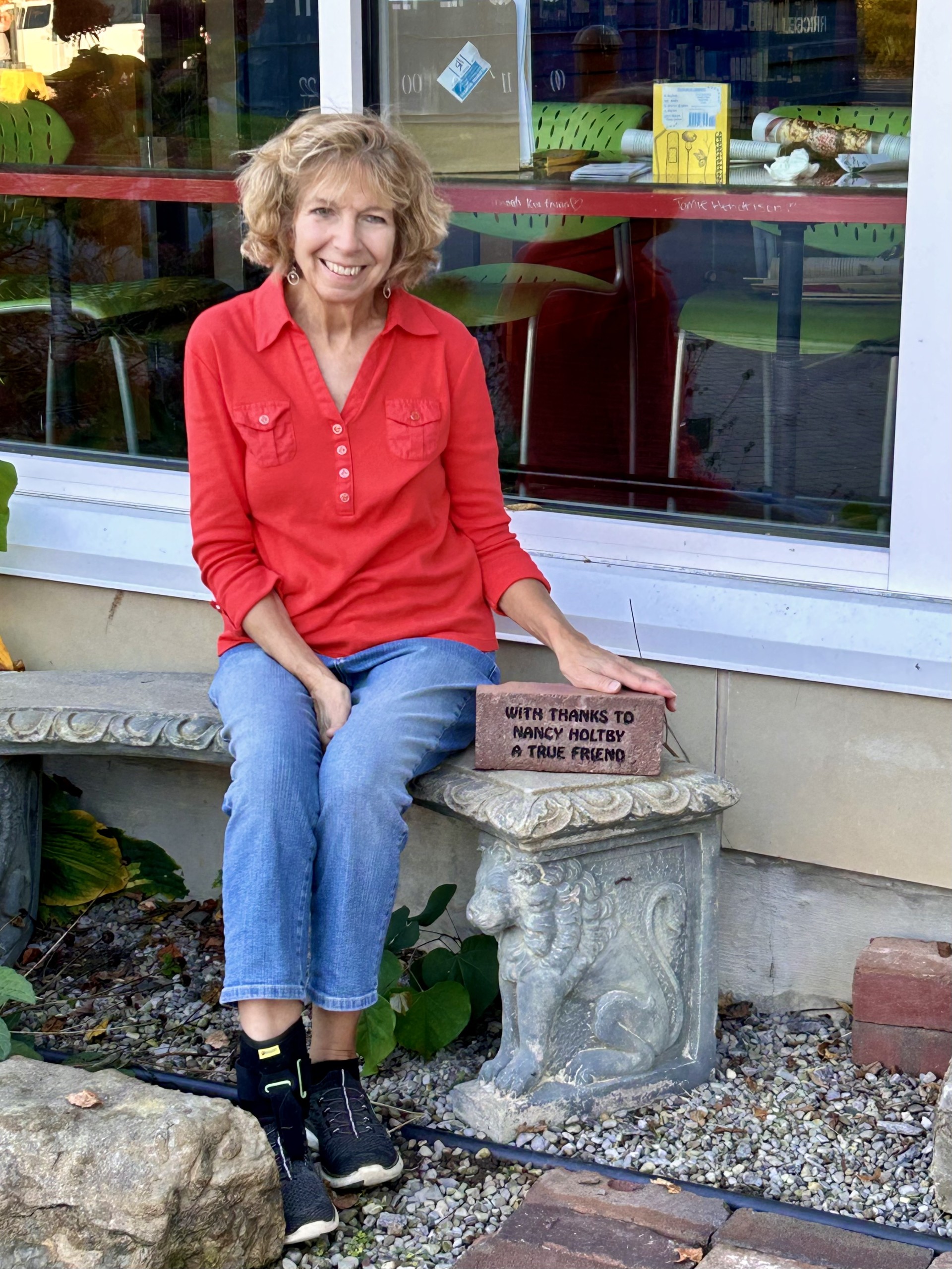 Nancy Holtby seated outside the Mendon Public Library next to the commemorative brick that was created in her honor for her dedicated time as President of The Friends of Mendon Public Library.